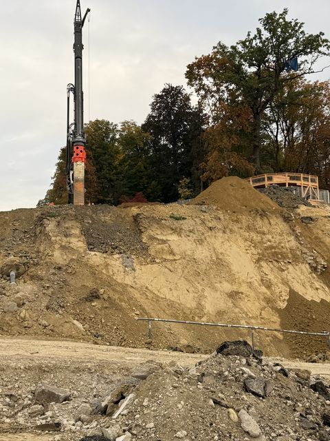 Ein abgegrabener Hang mit freigelegter Erde und Schutt, oben ein Bohrturm vor einer Baumreihe